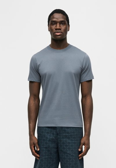 Άντρας που φοράει γκρι απλό κοντομάνικο μπλουζάκι και σκούρα μπλε καρό σορτς, στέκεται μπροστά σε μια απλή λευκή επιφάνεια.