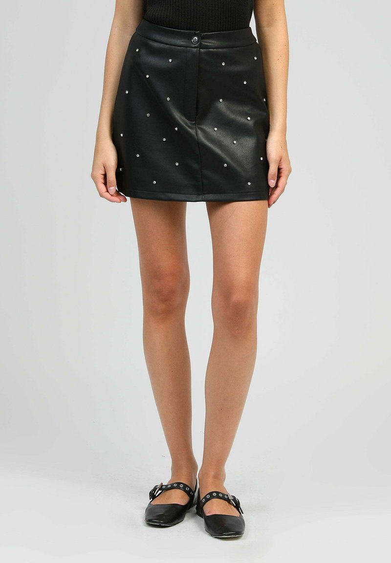 Femme portant une mini-jupe en cuir noire à clous, un haut en maille noire et des chaussures plates noires avec des boucles argentées, sur fond uni.