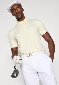 Polo shirt in lichtgeel stof met een kraag, voorzien van drie knopen. Gecombineerd met witte broek en een witte pet. Houdt een golfclub vast.