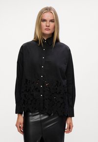 Camisa preta de botões com uma sobreposição de renda floral texturizada na barra, apresentando botões semelhantes a pérolas e mangas longas. Acabamento de tecido suave.