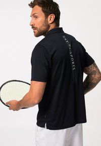 Polo en marine à manches courtes, avec un logo dans le dos et des fentes sur les côtés. Une silhouette masculine tient une raquette de tennis tout en se retournant.