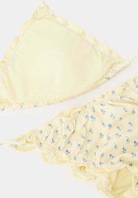 Bikini jaune avec motif floral bleu et bords festonnés, posé à plat sur un fond blanc.