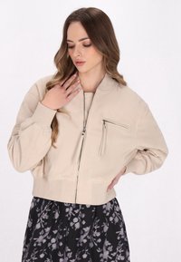 Giovane donna con lunghi capelli che indossa una giacca beige con zip e una gonna nera a fiori, in posa con una mano sul petto e l'altra sulla vita.