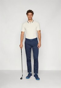 Ανδρική γκολφ εμφάνιση: κοντομάνικο λευκό πόλο με κόκκινο γιακά, σκούρες μπλε παντελόνες, σκούρα μπλε αθλητικά παπούτσια, κρατώντας ένα γκολφ κλαμπ, στέκεται σε απλό φόντο.