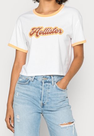 Kobieta ubrana w białą krótka koszulkę z pomarańczową lamówką i logo "Hollister", zestawioną z jasnoniebieskimi postarzanymi jeansami z wysokim stanem.