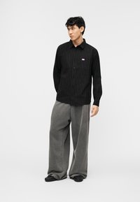 Νεαρός άνδρας που φοράει μαύρο πουκάμισο με ρίγες και φαρδιά γκρι παντελόνια, στέκεται με τα χέρια πίσω από την πλάτη πάνω σε λευκό φόντο.