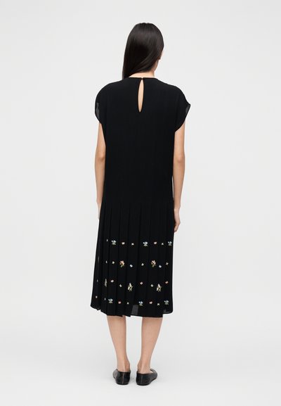 Robe longue noire à manches courtes, design plissé, dos échancré en forme de goutte, avec des motifs floraux brodés aux couleurs pastel près de l'ourlet.