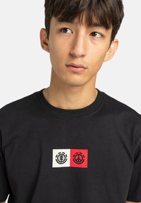 Zwarte katoenen t-shirt met een grafisch ontwerp met twee rechthoekige accenten: één wit met een boomsymbool, één rood met een vredessymbool.