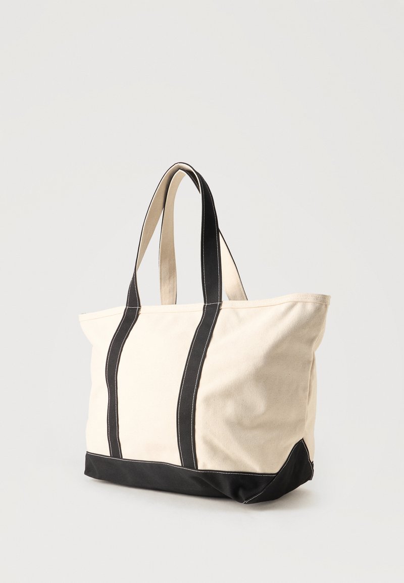 Canvas tote bag in beige with black straps and bottom, standing upright against a plain light background.