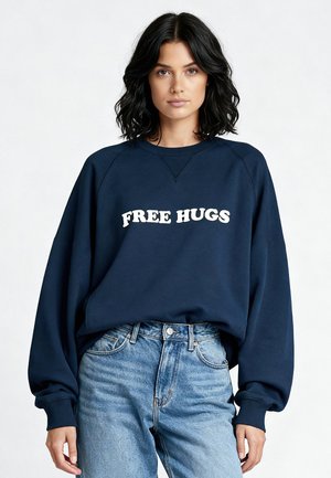 Felpa blu navy con maniche raglan e scritta bianca "FREE HUGS", abbinata a jeans blu chiaro a vita alta con vestibilità comoda.