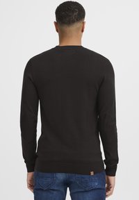 Donkerbruine trui met lange mouwen, geribde manchetten en zoom, met elleboogpatches. G gedragen met blauwe jeans. Soepele textuur.