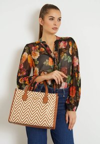 Håndtaske i tan og hvidt chevronmønster med to brune håndtag. Vævet tekstur, struktureret design. Model iført en blomsterbluse og denim.