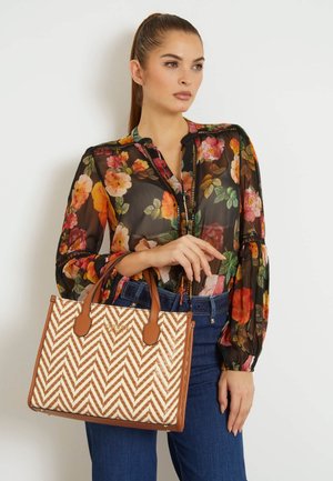 Tasche im Chevron-Muster in Sand- und Weißtönen mit zwei braunen Griffen. Geflochtene Textur, strukturierte Form. Modell trägt eine Blumenbluse und Jeans.