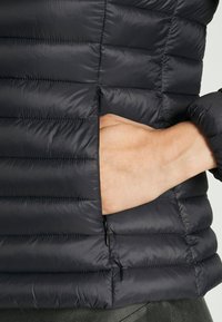 Zwarte puffer vest met een gladde, gewatteerde textuur, voorzien van een zijzak met rits en een aansluitend ontwerp rond de arm.
