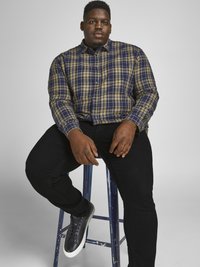 Plaid shirt in green, navy, and cream, buttoned up with a collar. Black jeans, seated on a blue stool, paired with dark sneakers.