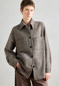 Brūna un bēša raiba jaka, kas izgatavota no teksturēta auduma, ar izteiktu apkakli, pogu aizdari un krūšu kabatu.