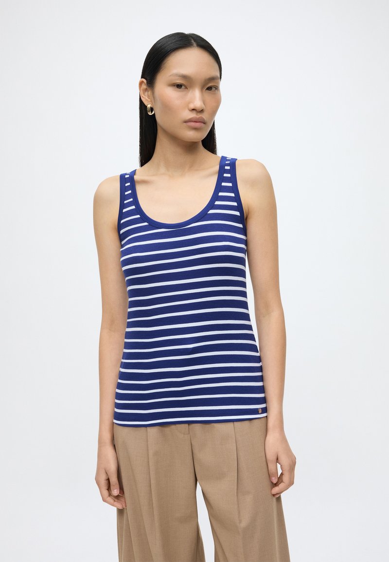 Femme aux cheveux noirs raides portant des boucles d'oreilles créoles en or, un débardeur rayé bleu et blanc et un pantalon beige sur fond blanc.