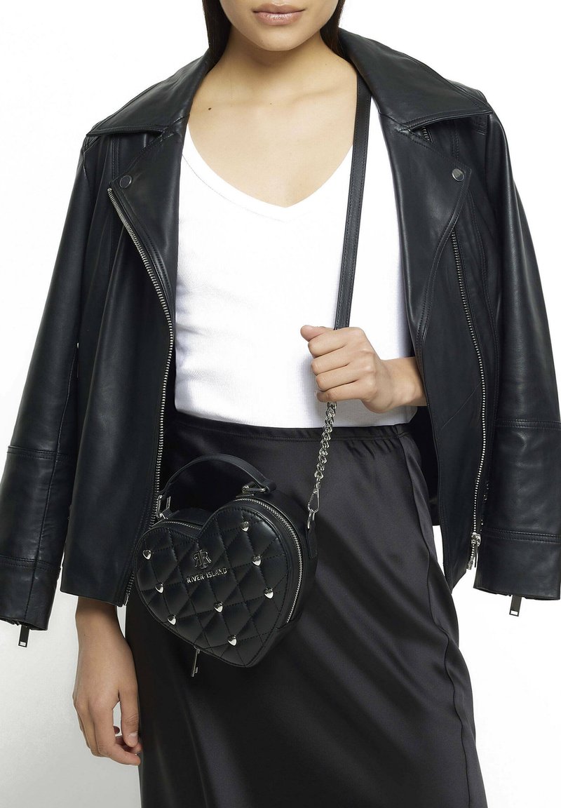 Černá kabelka ve tvaru srdce s prošívaným designem, stříbrnými cvočky a odnímatelným řetízkovým popruhem, v kombinaci s černou koženou bundou a bílým topem.