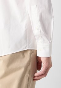 Camicia bianca a maniche lunghe con una texture liscia, caratterizzata da un unico polsino e un orlo dritto, abbinata a pantaloni khaki beige.