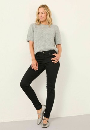 Frau mit blonden Haaren, die einen grauen kurzärmligen Pullover, schwarze enge Hosen und silberne Ballerinas trägt, steht vor einer weißen Wand.