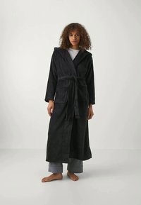 Zwarte badjas met capuchon van zacht materiaal, met een tailleband, lange mouwen en twee voorkeurszakken. Draag het over een grijze broek en een top.