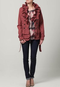 Bordeauxkleurige jas met een gerimpelde kraag, knoopsluiting aan de voorkant en zijvakken; gedragen over een gebloemde top, gecombineerd met donkere jeans en beige flats.