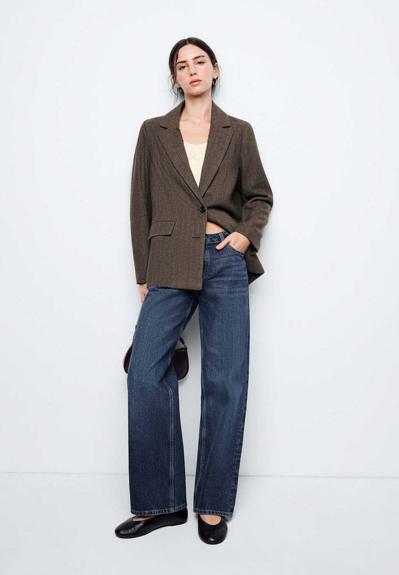 Femme debout contre un mur blanc, portant un blazer marron, un haut crème, un jean bleu à jambes larges et des chaussures noires, une main dans sa poche.