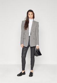 Juoda ir balta houndstooth stiliaus švarkas, prigludęs dizainas, derinamas su juodomis odinėmis kelnėmis ir loafers tipo bateliais, laikant juodą rankinę.