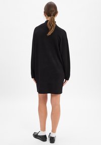 Czarna, oversize sukienka-sweter z wysokim kołnierzem, długimi rękawami, ściągaczami na mankietach i prostym dołem. Noszona z czarnymi butami i białymi skarpetkami za kostkę.