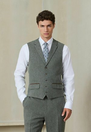 Jeune homme portant un gilet gris sur mesure, un pantalon assorti, une chemise blanche et une cravate à motifs, debout avec une main dans la poche.