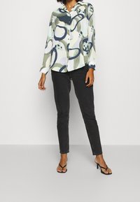 Donna in piedi con sicurezza, indossa una camicia a fantasia con bottoni e jeans neri, abbinati a sandali neri con tacco, su uno sfondo semplice.