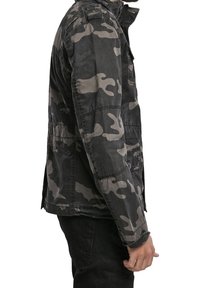 Kamouflagejacka i svart och grått, med ståkrage, flera fickor och ett strukturerat tyg. Sidsyn visar det lager på lager-design.