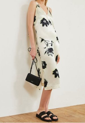 Cremefarbenes Slipkleid mit floralen Mustern in Schwarz, V-Ausschnitt und seitlichen Schlitzen. Kombiniert mit einer schwarzen Handtasche und flachen schwarzen Sandalen.