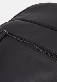 Sac à dos en cuir noir avec une surface texturée, une fermeture éclair centrale et un logo en relief indiquant "PIER ONE" à l'avant.