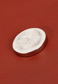 Bouton rond en métal argenté avec l’inscription en relief « THE ROW MADE IN ITALY », posé sur une surface en cuir texturé couleur rouille.