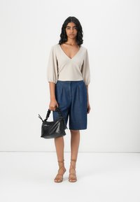 Top bege texturizado com um decote em V profundo, combinado com calções de ganga azuis. A segurar uma pequena mala preta com uma alça trançada. Sandálias de tiras em tom bronze.