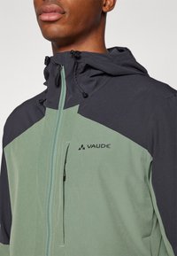 Veste zippée verte et noire en tissu léger, dotée d'une capuche, de cordons de serrage ajustables et d'un logo sur la poitrine.