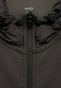 Gros plan sur la fermeture éclair d'une veste gris foncé avec un col rembourré et des cordons réglables de chaque côté, montrant l'étiquette de la marque MNG à l'intérieur du col.