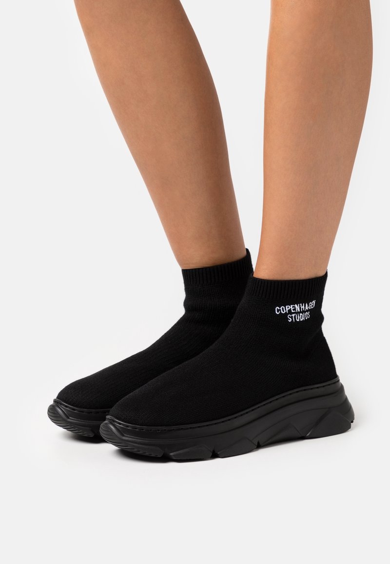 Jambes portant des baskets style chaussette noires à semelles épaisses, texte "Copenhagen Studios" sur la zone de la cheville de chaque chaussure.