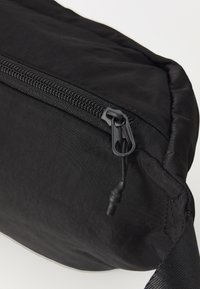 Black fabric shoulder bag with a textured surface, featuring a top zipper closure, slim design, and minimalist black strap.