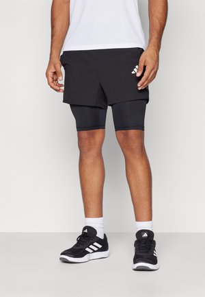 Mężczyzna ubrany w czarne warstwowe szorty sportowe, czarne szorty kompresyjne, białe skarpetki i czarne buty sportowe Adidas, stojący na jednolitym tle.
