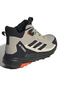 Botte de randonnée montante en gris clair avec des accents noirs, présentant un dessus texturé, une semelle noire, des détails orange et un col de cheville rembourré.