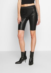 Pantalones cortos de motorista de piel sint�ética negra con un diseño ajustado, de longitud a mitad del muslo y una textura suave. Usados con botas negras hasta el tobillo.