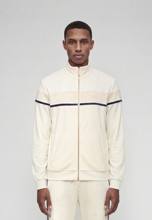 Homme portant une veste de survêtement crème zippée avec des rayures horizontales beige et noir, debout, les bras détendus le long du corps.