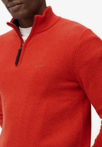 Czerwony sweter z półkola z zamkiem błyskawicznym, dzianinowa faktura i małe logo na piersi. Wykonany z miękkiego, oddychającego materiału.