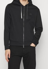 Uomo che indossa una felpa nera con zip e cordini bianchi e neri, pantaloni neri, con una mano in tasca, su uno sfondo semplice chiaro.