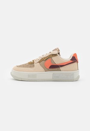 Niskie buty sportowe z zamszu w beżowym i jasnobrązowym kolorze, z pomarańczowym i ciemnoczerwonym logo Nike oraz białą gumową podeszwą.