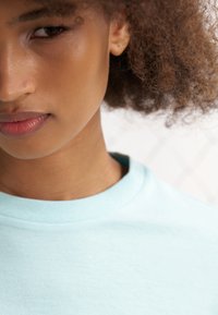 Menta zöld pamut póló kerek nyakkivágással, enyhén texturált felülettel és látható varrási részletekkel. A háttér elmosódott.