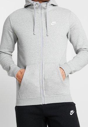 Mężczyzna w jasnoszarej bluzie Nike na zamek z kieszeniami i czarnych spodniach Nike, ręce w kieszeniach bluzy, zwrócony przodem.
