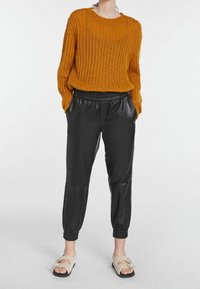 Pomarańczowy sweter z dzianiny o luźnym kroju, w połączeniu z czarnymi dresami z materiału przypominającego skórę, z elastycznym paskiem w talii i mankietami na kostkach. Sportowe lekkie sandały.